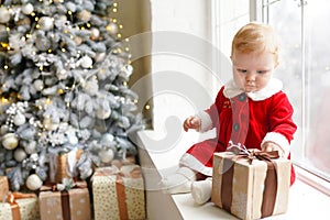 Positive photo of cute beautiful little girl in Santa`s dress playing christmas gift box on the windowsill