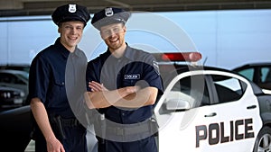 Positive police mates in hats smiling into camera against squad car, work