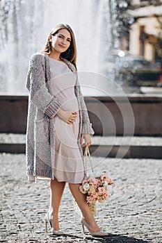 Positive pregnant girl with a bouquet of flowers walking near the fountain. Fine weather