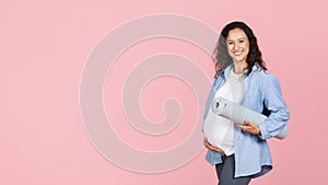 Positive pregnant woman holding fitness mat, pink background