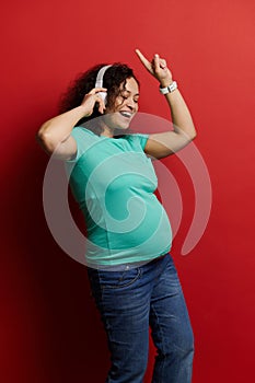 Positive pregnant woman wearing wireless headphones, having fun, dancing, listening to music, isolated on red background