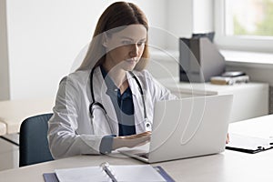 Positive pretty doctor woman typing on laptop at work table