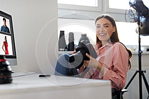 Positive pretty lady photographer working in studio, sitting at workdesk with computer, holding professional camera