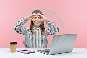 Positive pretty woman showing heart gesture with hands sitting at workplace with laptop, smiling expressing romantic amorous