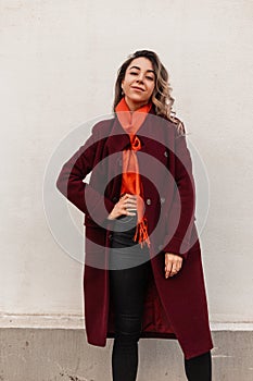 Positive pretty young woman fashion model with vintage orange scarf in stylish burgundy coat posing near vintage wall. Fashionable