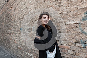 Positive pretty young woman in a stylish black T-shirt in a luxurious long coat in white jeans in a beautiful smile