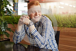 A positive redhead hipster with freckles wearing stylish checked shirt and watch sitting at wooden table in cosy restaurant having