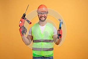 Positive repairman holds hammer and drill,wears yellow protective construction helmet, reflective waistcoat and protective glasses