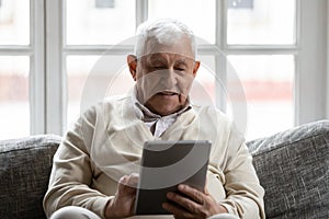 Positive retired man relaxing on comfy couch using tablet computer