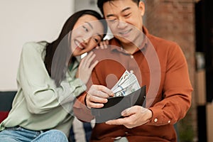 Positive rich middle aged asian man putting banknotes into wallet, sitting with his young wife on sofa at home