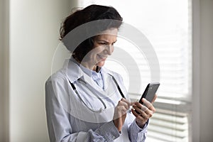 Positive satisfied mature head doctor woman using smartphone in office