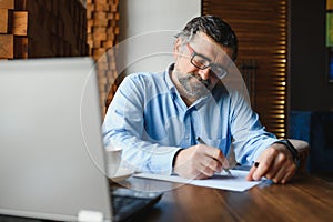 Positive senior bearded man with grey hair drinking coffee and using laptop at cafe, copy space. Stylish aged