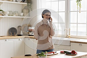 Positive senior chef woman speaking on cellphone in home kitchen