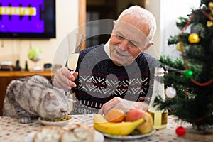 Positive senior man sitting at home table with cat during Christmas celebration