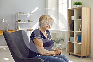 Positive senior retired woman grandmother in jeans and t-shirt sitting with glass of water in armchair