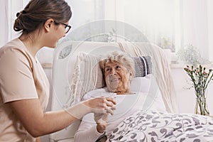 Positive senior woman lying in bed, helpful doctor in beige uniform supporting her