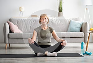 Positive senior woman sitting in lotus pose on sports mat, meditating at home, copy space