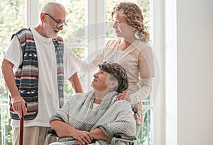 Positive senior woman on wheelchair with caring nurse and elderly friend with walking stick