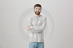 Positive shy good-looking guy with beard standing with crossed hands in pullover over gray background, smiling and