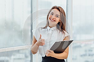 Positive smiling female teacher inspired with education ideas, stands holding paperholder, giving thumb up.