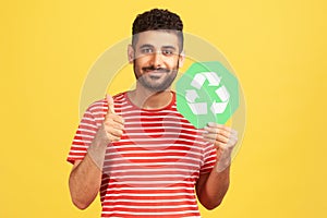 Positive smiling man with beard showing thumbs up gesture holding green waste recycling symbol, satisfied with environmental