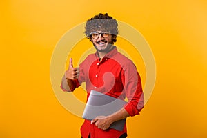Positive smiling man freelancer with beard showing thumbs up while standing and holding laptop, satisfied with