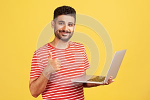 Positive smiling man freelancer with beard in striped t-shirt showing thumbs up standing and holding laptop, satisfied with