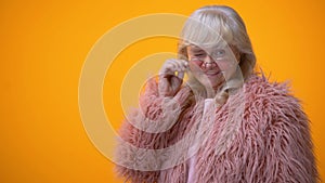 Positive smiling senior lady in pink coat and round sunglasses winking, flirt