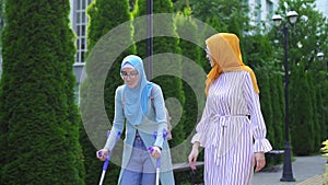 Positive smiling young woman in hijab on crutches after injury with girlfriend walking in Park