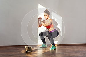 Positive sportive woman with bun hairstyle and in tight sportswear doing squatting. indoor studio shot illuminated by sunlight