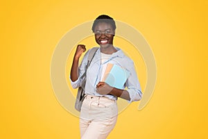 Positive teen african american lady student with books, exuding triumph and joy, raises fist