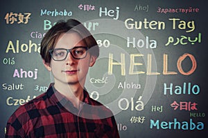 Positive teen guy wearing red shirt and eyeglasses over grey wall background and the word hello written in different languages and