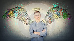 Positive teenage student boy keeps hands folded, tooth smile imagine himself an angel with fluffy wings behind back and a halo