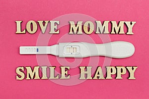 A positive test for pregnancy and the words `love, mommy, smile, happy` made of wooden letters on a pink background