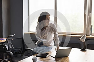 Positive thoughtful office employee girl sitting on table with laptop