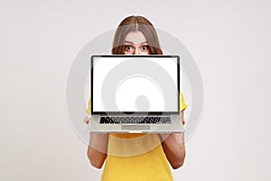 Positive unknown female in yellow T-shirt hiding half of face behind laptop with white empty display