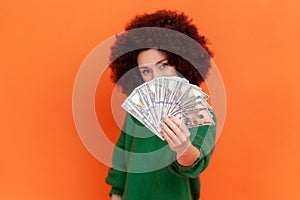 Positive woman with Afro hairstyle wearing green casual style sweater holding out fan of dollars