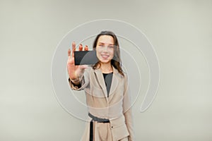 Positive woman in a beige suit shows a black bank card to the camera on a beige background. Copy space. Lady and debit card