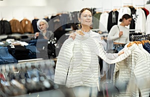 Positive woman chooses white winter down jacket in clothing store