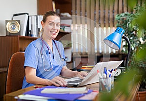 Positive woman doctor working in cabinet on laptop