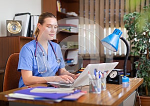 Positive woman doctor working in cabinet on laptop