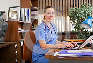 Positive woman doctor working in cabinet on laptop