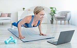 Positive woman doing plank on mat with laptop at home