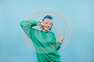 Positive woman in green sweatshirt and with colored hair dances on a blue background with a smile on her face