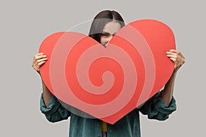 Positive woman hiding face behind big red heart and looking at camera, feeling affection fondness. photo