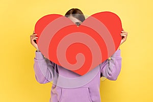 Positive woman hiding her face behind big red heart and looking at camera with curious prying eyes.