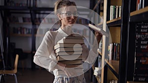 Positive woman holding pile of books in the library, putting more on the stack