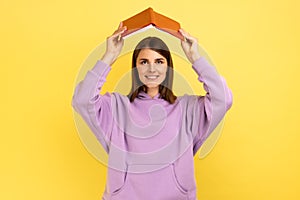 Positive woman making roof, holding book over head, looking at camera, expressing happiness.