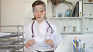 Positive woman physician sitting at table in her office and using laptop.