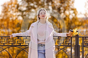 Positive woman is sitting in the park with takeaway coffee. Autumn